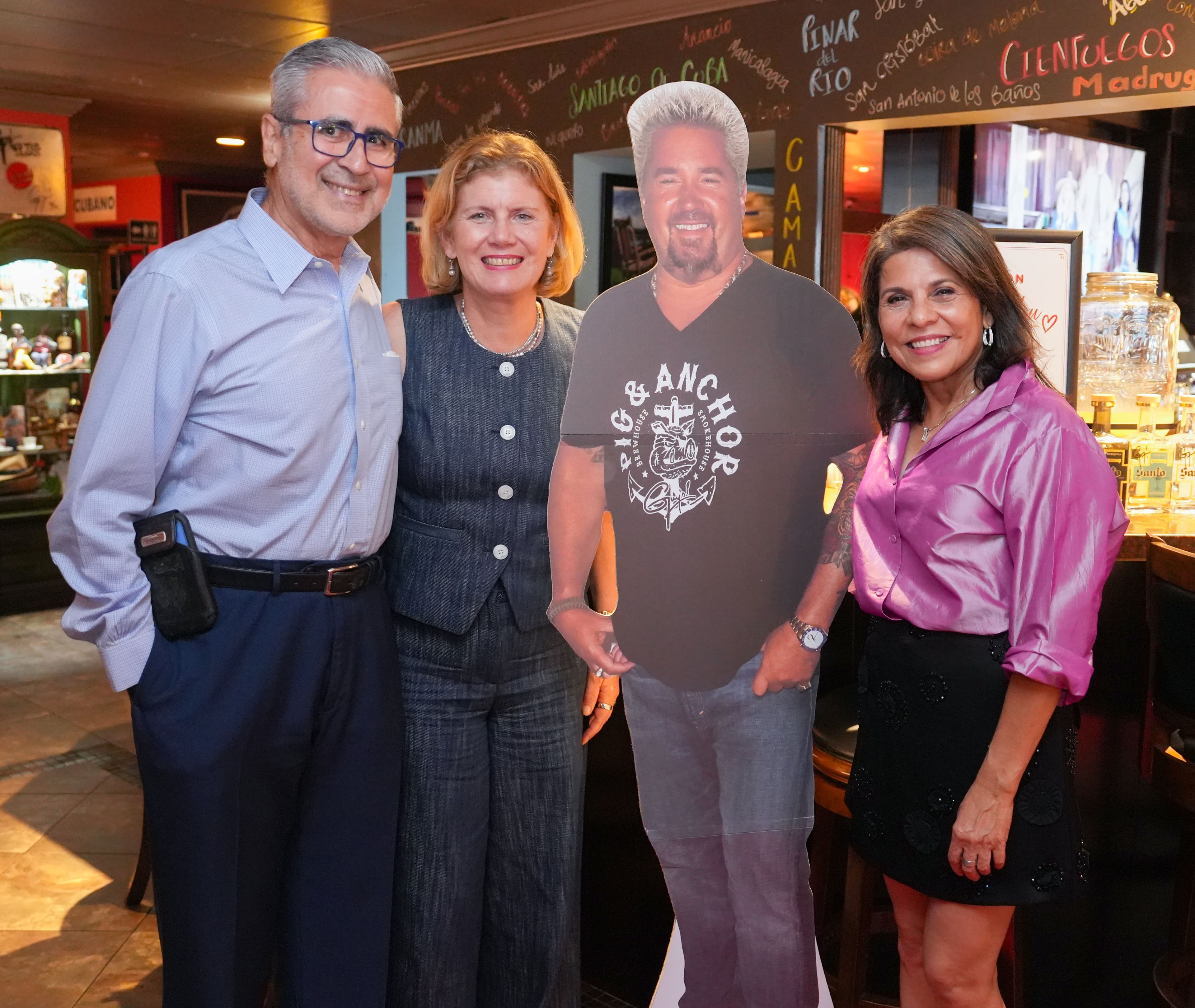 Three guests beside the Guy Fieri cutout under the chalkboard mural of Cuban town names