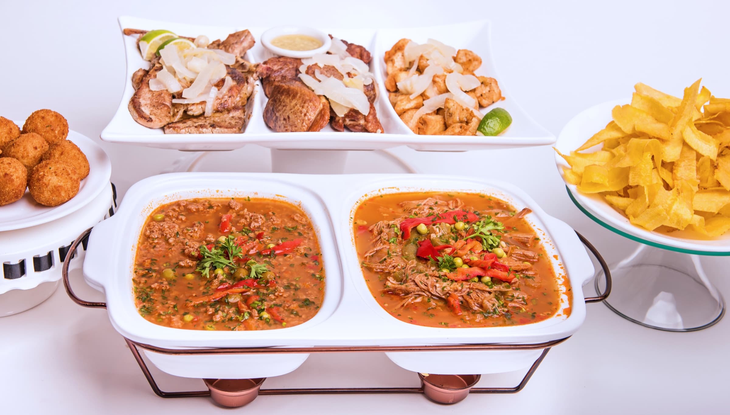 A catering buffet: picadillo and ropa vieja in a chafing dish, a tray of pork, steak and chicken chunks with onions, croquetas, and a stand of plantain chips